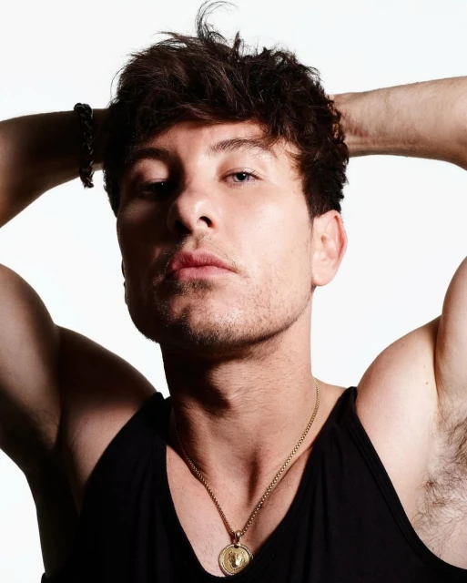 Portrait of a young adult man with dark hair, wearing a black tank top and a gold chain, posing with his hands behind his head.