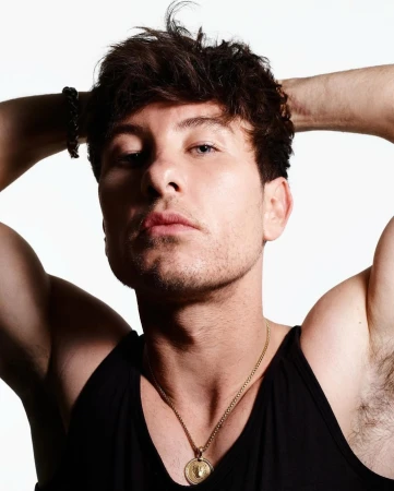 Portrait of a young adult man with dark hair, wearing a black tank top and a gold chain, posing with his hands behind his head.
