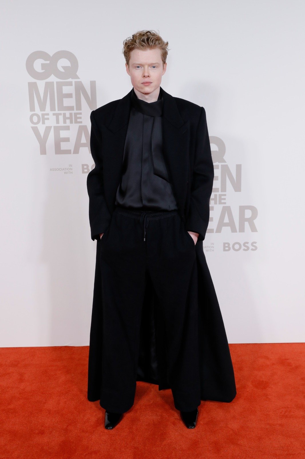 Man in a black overcoat and black suit stands on a red carpet in front of a GQ Men of the Year backdrop, looking straight at the camera.