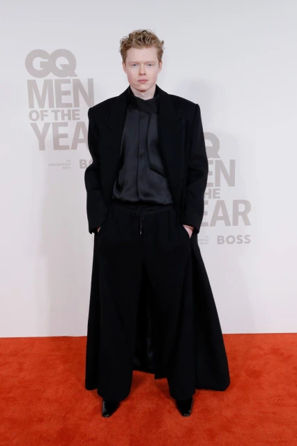 Man in a black overcoat and black suit stands on a red carpet in front of a GQ Men of the Year backdrop, looking straight at the camera.