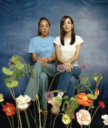 Two women seated against a mottled blue backdrop with colorful flowers in the foreground, styled by Charley Mcewen