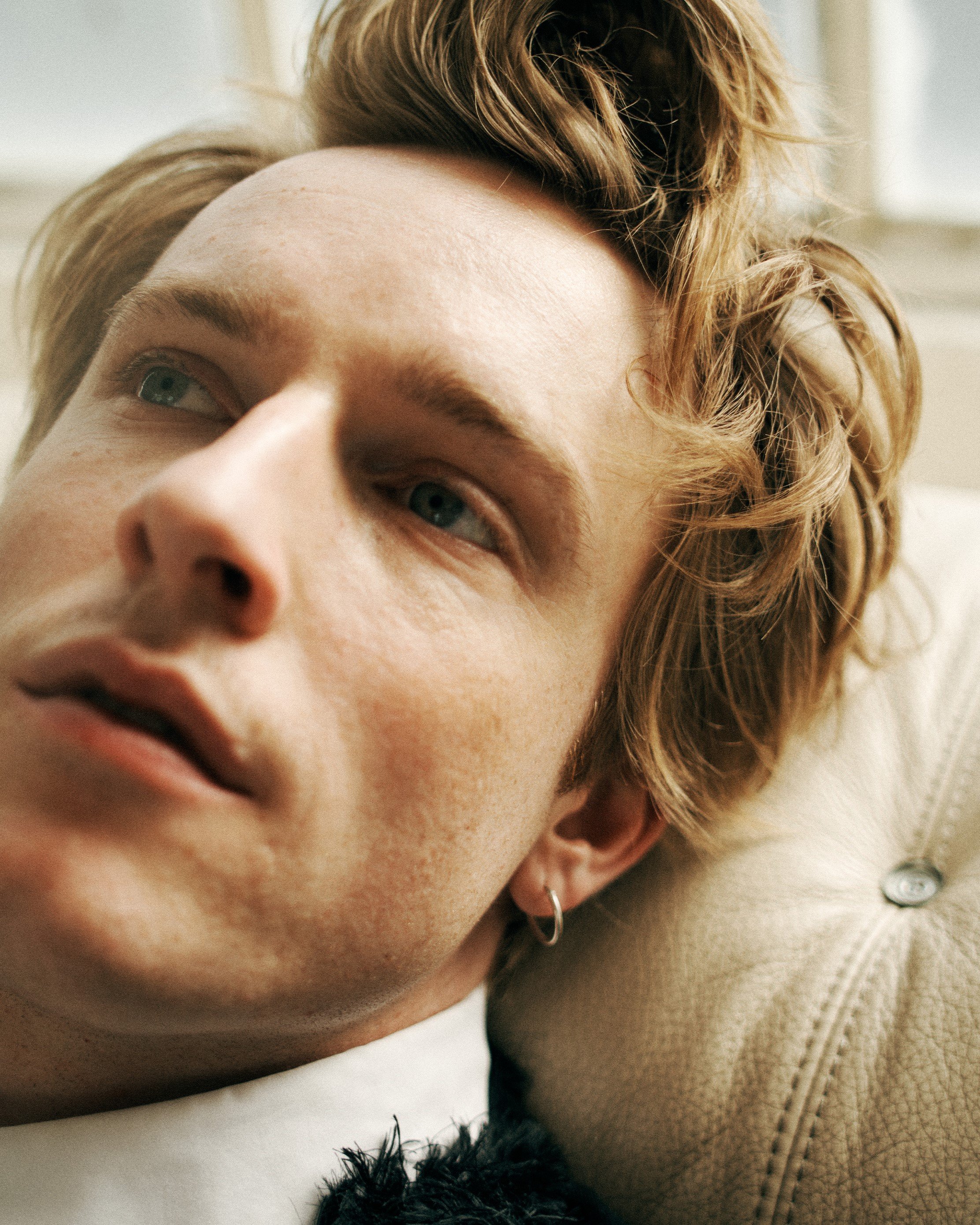 Close-up portrait of a young man with blonde hair and blue eyes, wearing a white shirt and silver hoop earring, leaning against a textured cushion.