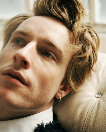 Close-up portrait of a young man with blonde hair and blue eyes, wearing a white shirt and silver hoop earring, leaning against a textured cushion.