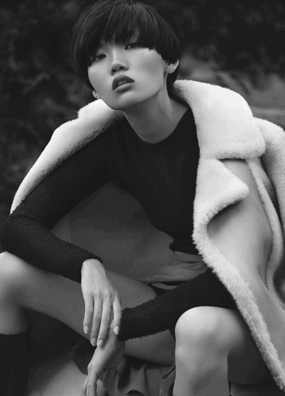 An Asian woman with short dark hair poses outdoors in a black long-sleeved top, tan shorts, and a white coat. Black and white.