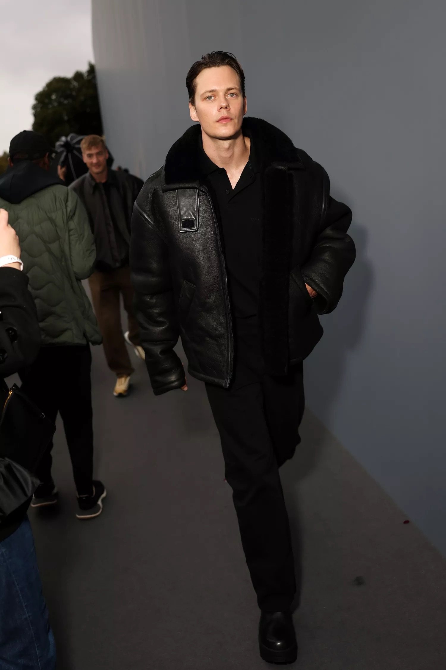 Adult male with fair skin and dark hair walks wearing an aviator-style jacket and black shoes with other people in the background.