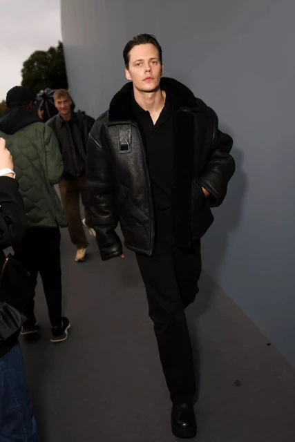 Adult male with fair skin and dark hair walks wearing an aviator-style jacket and black shoes with other people in the background.