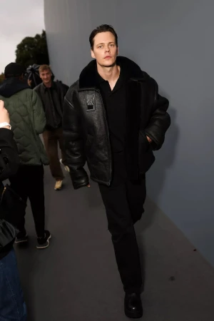 Adult male with fair skin and dark hair walks wearing an aviator-style jacket and black shoes with other people in the background.