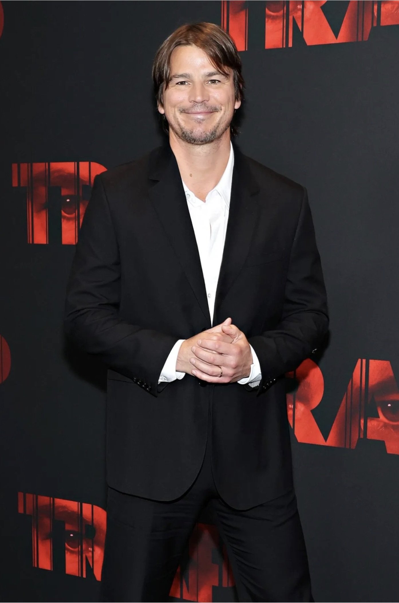 Josh Holloway poses in a black suit and white shirt against a red and black backdrop, looking confidently at the camera, medium shot.