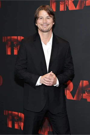 Josh Holloway poses in a black suit and white shirt against a red and black backdrop, looking confidently at the camera, medium shot.