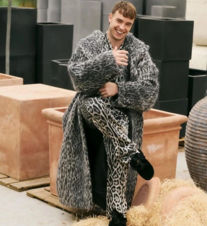 Smiling man wearing a leopard print coat and pants sitting in terra cotta planters in an outdoor setting with black planters behind.