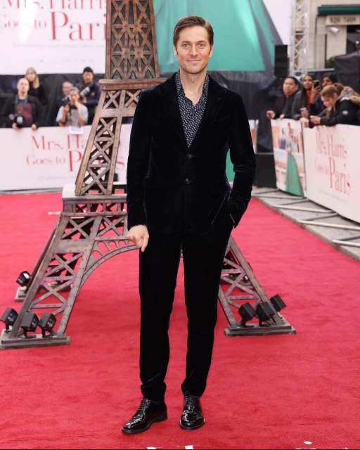 Man in a velvet suit posing on a red carpet with an Eiffel Tower replica, promoting the film 'Mrs. Harris Goes to Paris.'