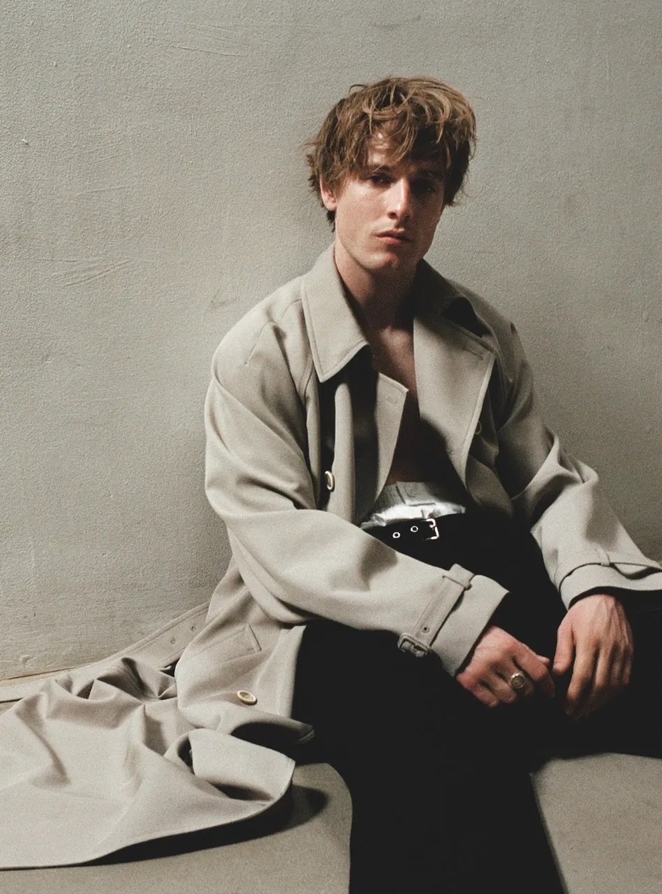Man in unbuttoned trench coat sitting on floor, looking directly at camera in neutral indoor studio setting with blank background.