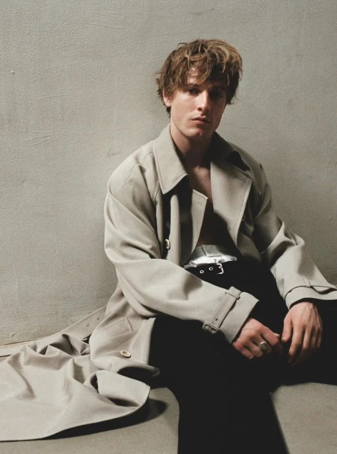 Man in unbuttoned trench coat sitting on floor, looking directly at camera in neutral indoor studio setting with blank background.