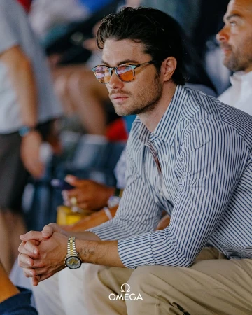 Portrait of a dark-haired adult male wearing glasses and a striped shirt, seated outdoors, with a watch visible on his wrist.