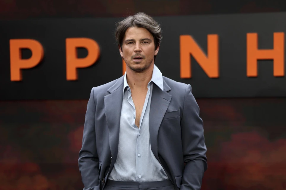 Adult man wearing a blue-grey suit and a light blue shirt with a partially open collar, posing against a backdrop of orange text.