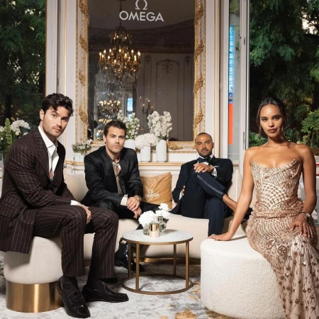 Group of four people, including two men in suits sitting on a white couch with an elegant room and a woman in a gown.