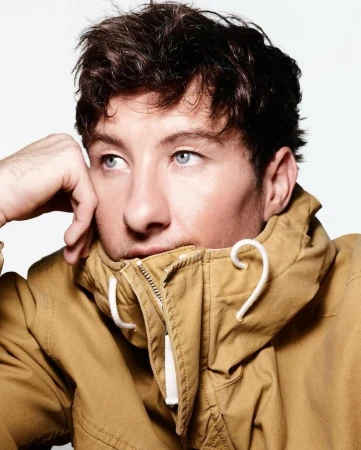 Close-up portrait of Barry Keoghan wearing a tan jacket with his hand on his cheek against a plain white background