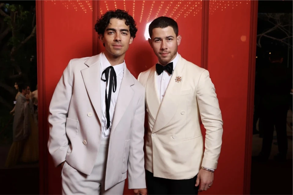 Two adult men in off-white suits pose against a red backdrop with subtle patterns, suggesting a formal event or premiere setting.