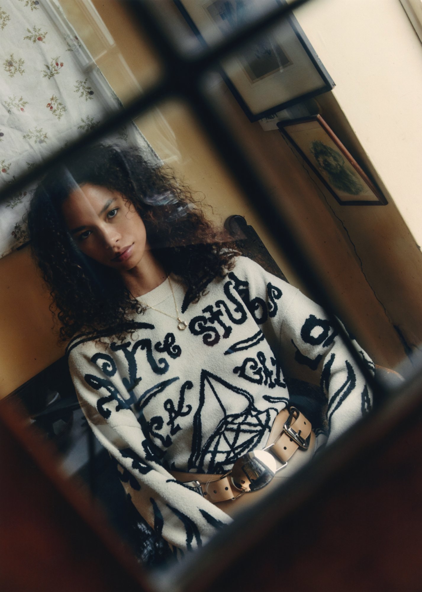 A young woman with curly hair poses indoors, wearing a patterned sweater and wide belt, reflected in a mirror with window frame.
