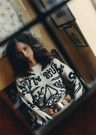 A young woman with curly hair poses indoors, wearing a patterned sweater and wide belt, reflected in a mirror with window frame.