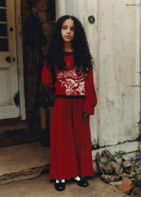 Young girl with long dark curly hair stands near a rustic white door, wearing a red sweater and long red skirt with black shoes.