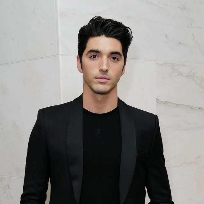 Portrait of a man with dark hair wearing a black blazer and t-shirt against a light marble background