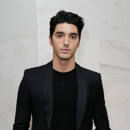 Portrait of a man with dark hair wearing a black blazer and t-shirt against a light marble background