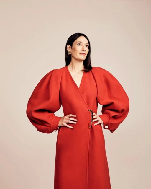 A woman stands confidently in a red wrap dress with large sleeves against a soft pink background, gazing upwards with slight smile.