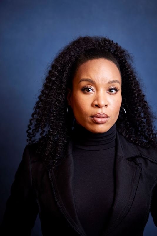 Portrait of a Black woman with long dark curly hair wearing a black turtleneck and blazer against a textured blue backdrop
