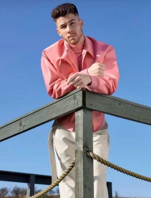 Portrait of a man wearing a pink shirt leaning on a wooden railing, set against a bright blue sky in an outdoor setting.