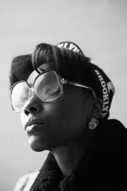 Black and white portrait of an African American woman with vintage 70s-style glasses, a patterned headscarf, and dark coat.