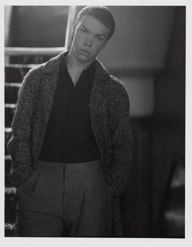 Black and white portrait of a fair-skinned young man in a dark shirt and speckled coat, standing in front of stairs.