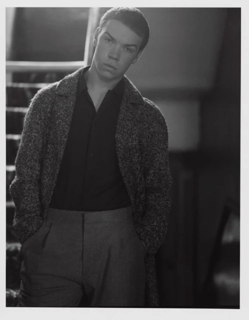 Black and white portrait of a fair-skinned young man in a dark shirt and speckled coat, standing in front of stairs.