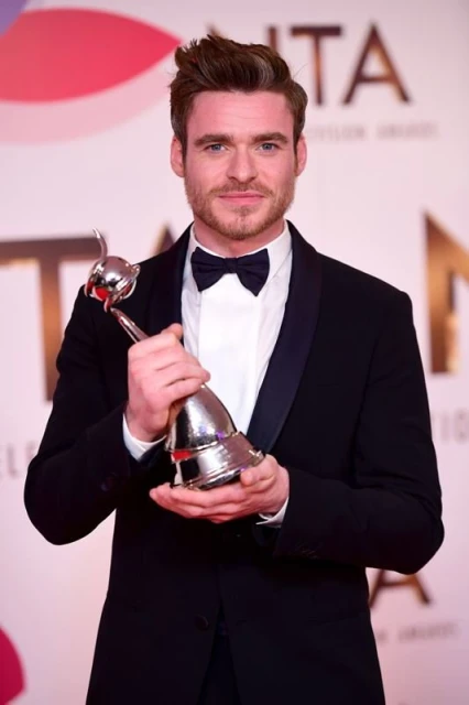 Adult man in black tuxedo smiles holding an award in front of a branded backdrop at a formal event, illuminated by photo studio lighting.
