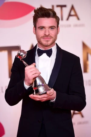 Adult man in black tuxedo smiles holding an award in front of a branded backdrop at a formal event, illuminated by photo studio lighting.