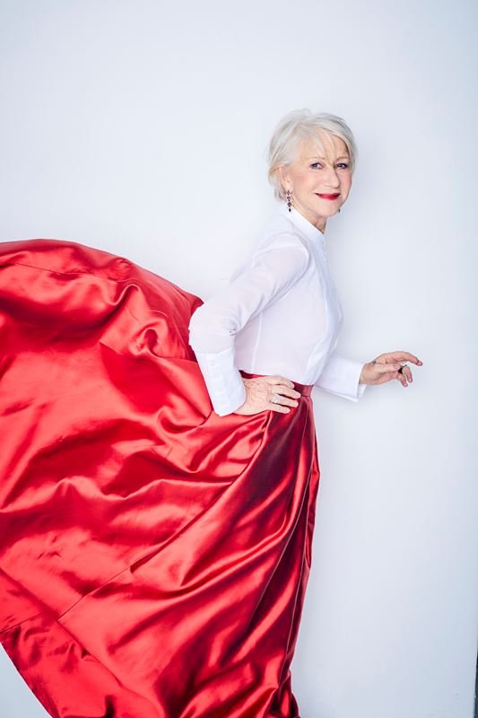 Helen Mirren in a red, flowing skirt and white blouse stands against a white background in a studio setting, smiling for the camera.