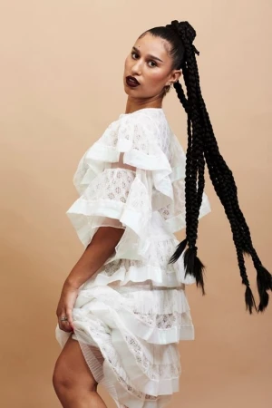 A woman with long braids, wearing a white, tiered, lace dress with ruffles poses against a neutral backdrop in a fashion shot.