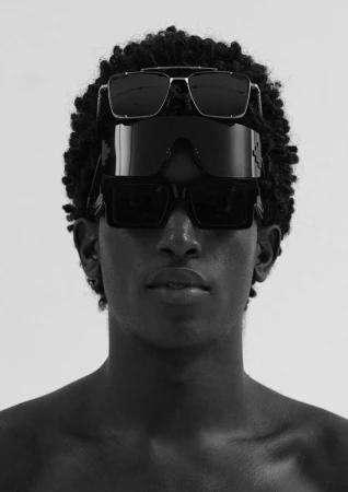 Close-up portrait of a Black man wearing three stacked pairs of dark sunglasses on his head and face against a light background.