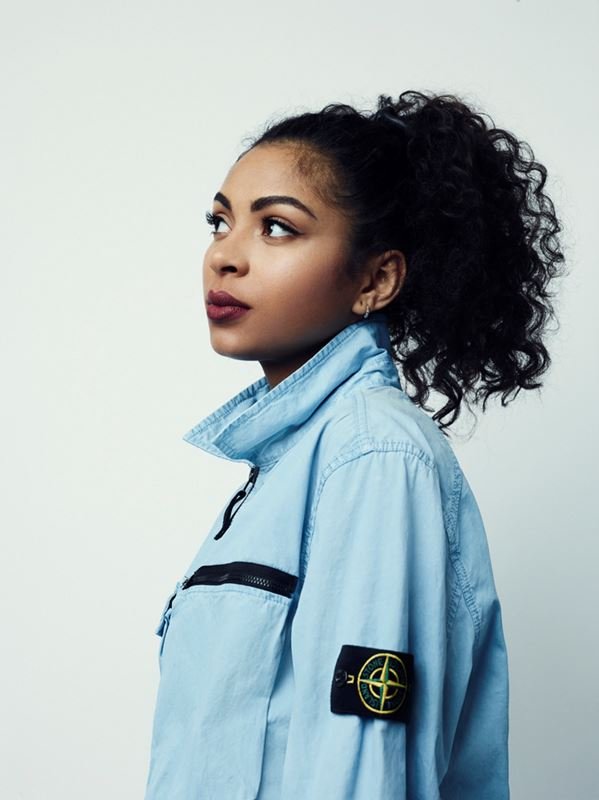 Profile portrait of a young adult woman with dark curly hair in a ponytail, wearing a light blue jacket against a white background.