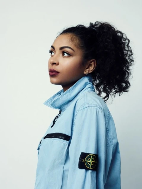Profile portrait of a young adult woman with dark curly hair in a ponytail, wearing a light blue jacket against a white background.