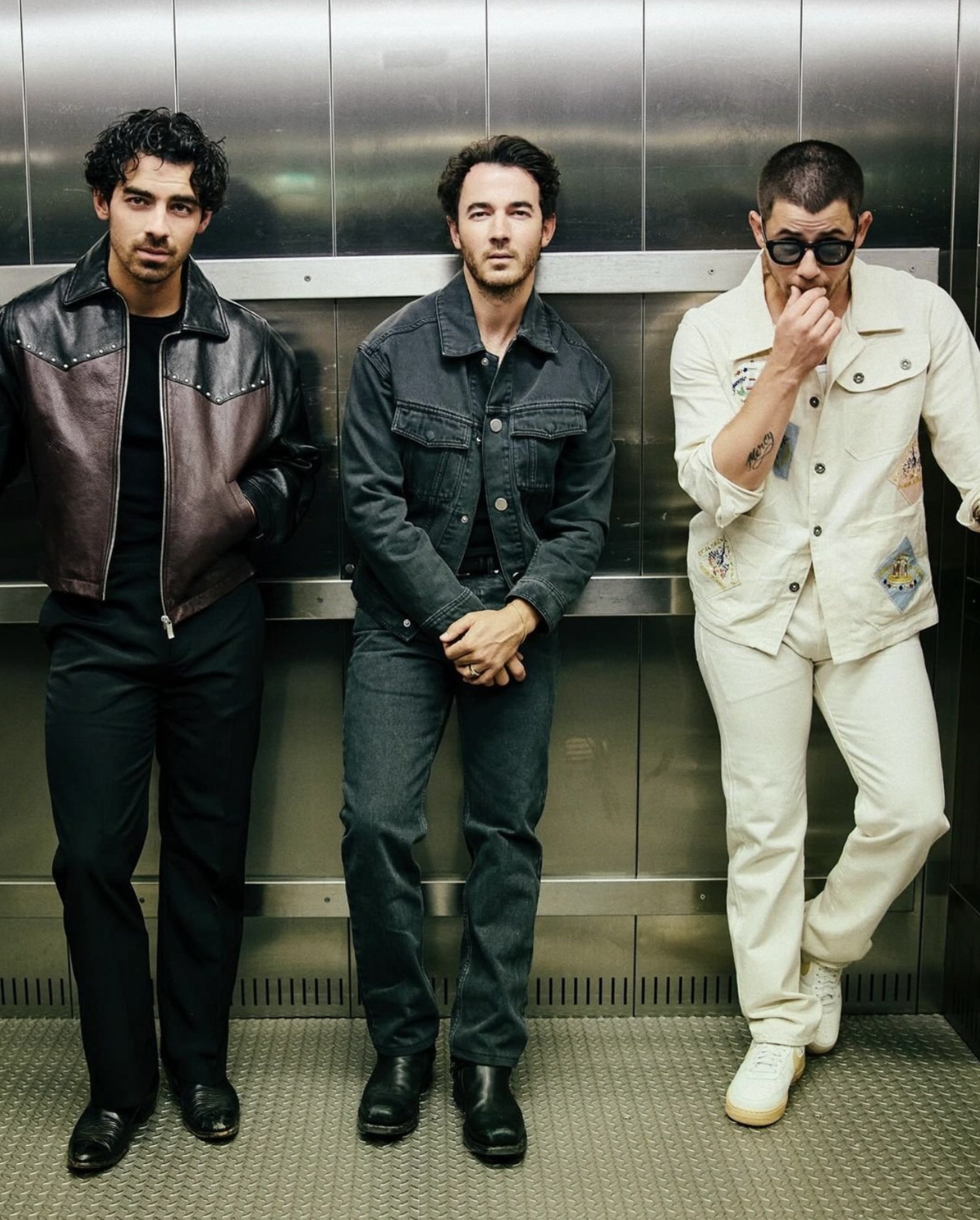 Three adult males stand in an elevator wearing casual jackets, jeans, and shoes; indoor setting with metal walls.