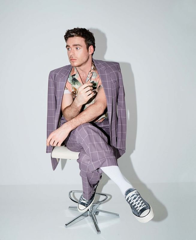 Man in plaid suit and floral shirt sitting on a stool in photo studio. Full shot of Richard Madden, indoor studio lighting, neutral backdrop.