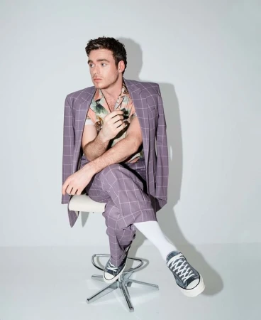 Man in plaid suit and floral shirt sitting on a stool in photo studio. Full shot of Richard Madden, indoor studio lighting, neutral backdrop.