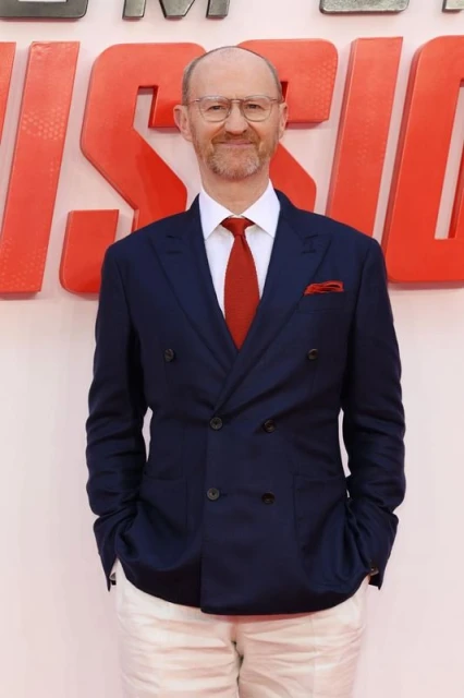 A man with glasses in a navy suit and white pants stands on a red carpet with a smiling expression, at an entertainment event.