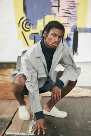 Young Black man with braided hair crouching in a light grey jacket and dark turtleneck against an abstract painted backdrop.