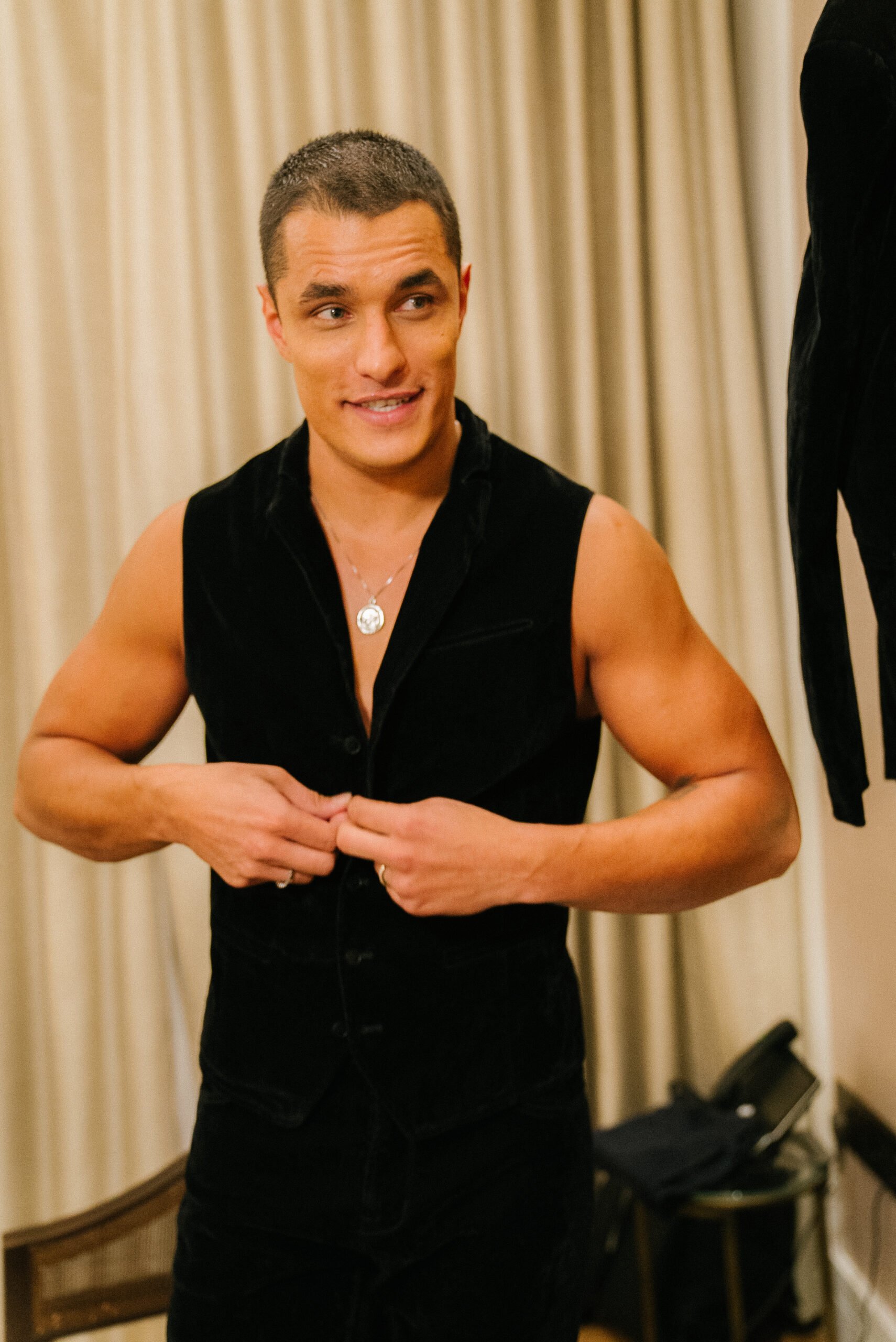 Attractive adult male in black vest buttons garment in indoor setting with soft lighting against neutral background, posing for portrait.