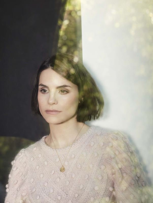Portrait of a woman with short brunette hair and a cream-colored sweater, gazing off-camera, posing in front of a blurred background.