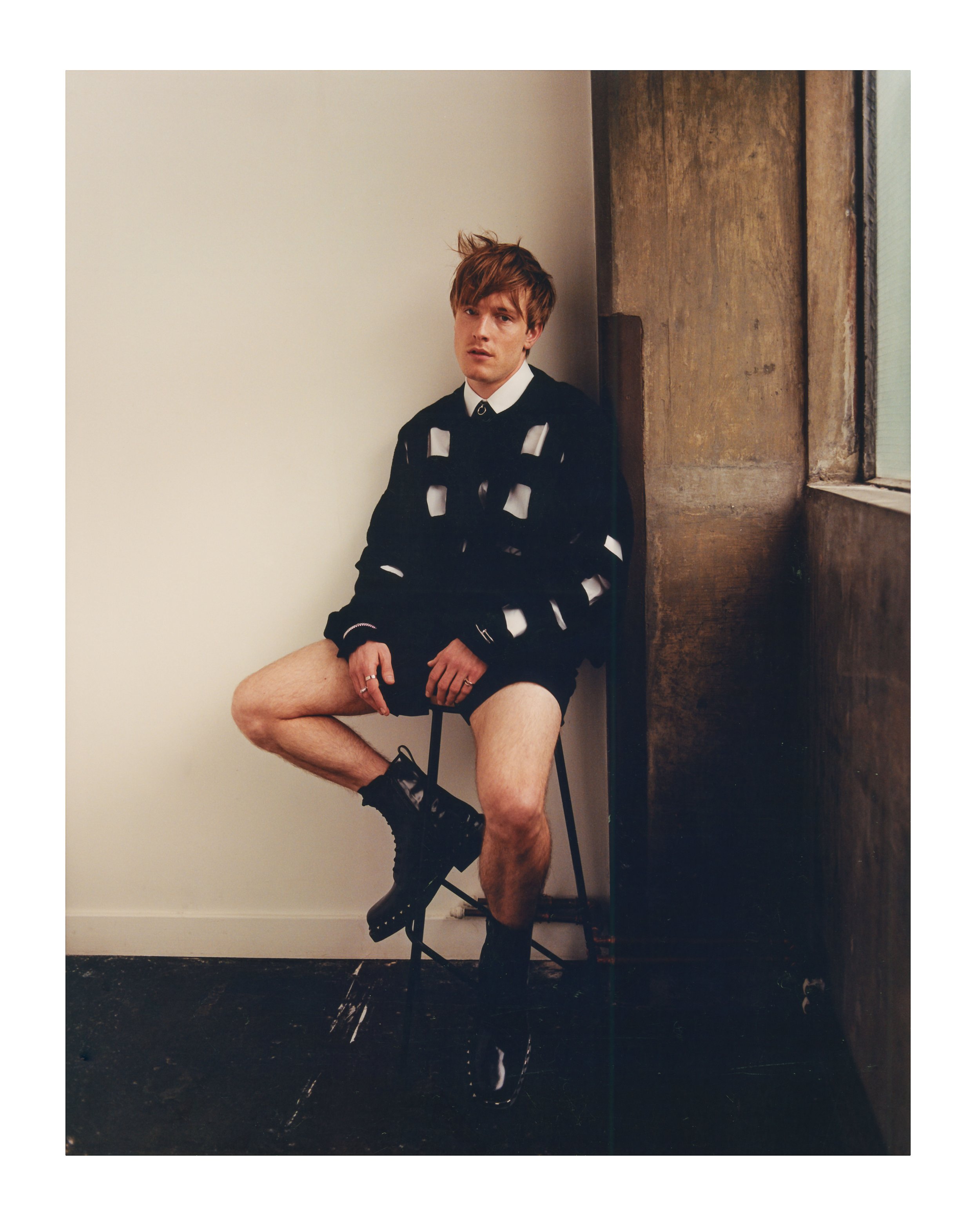 Young man with reddish-brown hair seated on a stool wearing a black cut-out knit sweater and black boots against a plain wall.