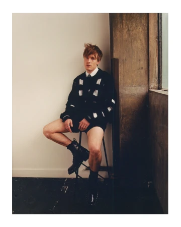 Young man with reddish-brown hair seated on a stool wearing a black cut-out knit sweater and black boots against a plain wall.