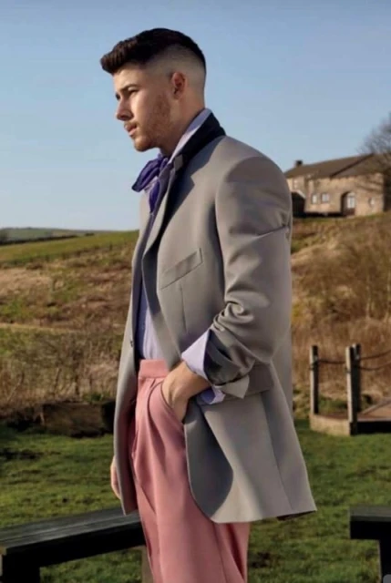 Portrait of a man in a grey suit and pink pants standing outdoors with a farmhouse and green fields in the background.